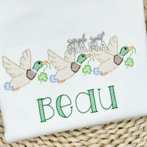 May include: Embroidered design of three ducks with shamrocks in their beaks, with the name "Beau" stitched below. The design is on a white fabric background.