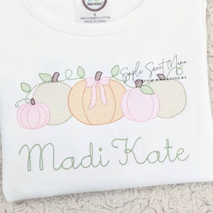 May include: A white t-shirt with an embroidered design of four pumpkins with pink and green accents. The text "Simple Sweet Mama Custom Embroidery" and the name "Madi Kate" are also embroidered on the shirt.