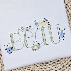 May include: A white embroidered shirt with the name "Beau" in green outlined letters. The letters are decorated with green and yellow bugs and a green grasshopper.