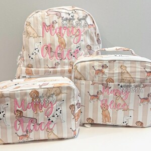 May include: A set of three matching bags: a backpack, a lunch bag, and a cosmetic bag. The bags feature a cream and tan striped pattern with various dog illustrations. The name "Mary Alice" is embroidered in pink.