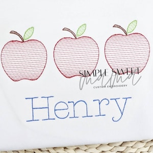 May include: Embroidered design of three red apples with green leaves and the name "Henry" in blue thread on a white fabric background.