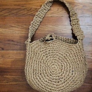 May include: A tan crocheted tote bag with a circular base and a long strap. The bag is made of natural fibres and has a rustic, handmade look.