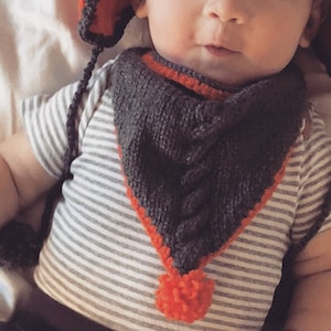 Puede incluir: Conjunto de gorro y babero de punto gris con ribete naranja y pompón. El babero tiene un diseño de punto trenzado y se lleva sobre una camiseta de rayas grises y blancas. El gorro tiene orejeras y lazos.