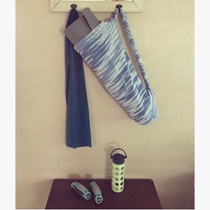 May include: A blue and white yoga mat carrier with a yoga mat inside, hanging on a hook. A blue towel is hanging on a hook next to it. A green water bottle and two black hand weights are on a wooden surface.