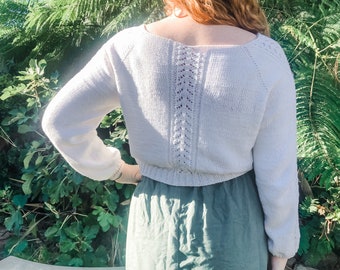 May include: A cream-coloured, long-sleeved jumper with a boat neck and a decorative openwork design down the back. The sleeves have a slight puff and are cuffed at the wrists. The jumper is paired with a sage green skirt.