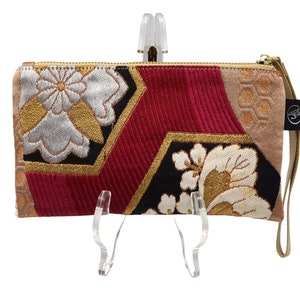 K&ouml;nnte beinhalten: Eine Clutch mit rotem, schwarzem, goldenem und wei&szlig;em Muster, einem goldenen Rei&szlig;verschluss und einem braunen Riemen. Die Clutch hat ein geometrisches Design mit floralen Akzenten.