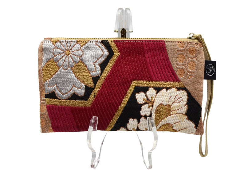 K&ouml;nnte beinhalten: Eine Clutch mit rotem, schwarzem, goldenem und wei&szlig;em Muster, einem goldenen Rei&szlig;verschluss und einem braunen Riemen. Die Clutch hat ein geometrisches Design mit floralen Akzenten.