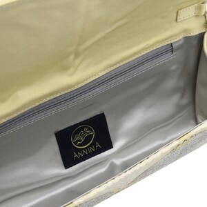 May include: A light yellow clutch bag with a silver chain strap and a silver and white patterned design. The bag has a zippered pocket inside and a black label with the word "ANNINA" on it.