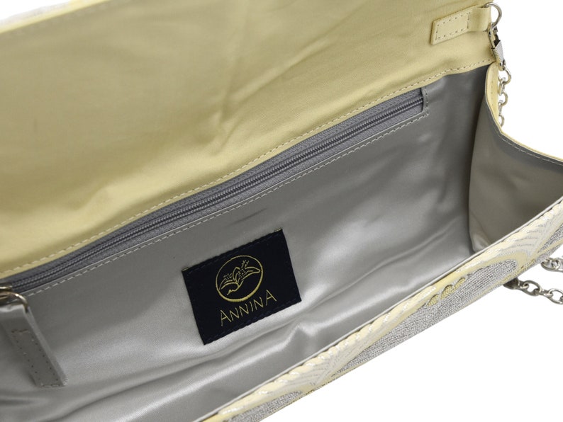 May include: A light yellow clutch bag with a silver chain strap and a silver and white patterned design. The bag has a zippered pocket inside and a black label with the word "ANNINA" on it.