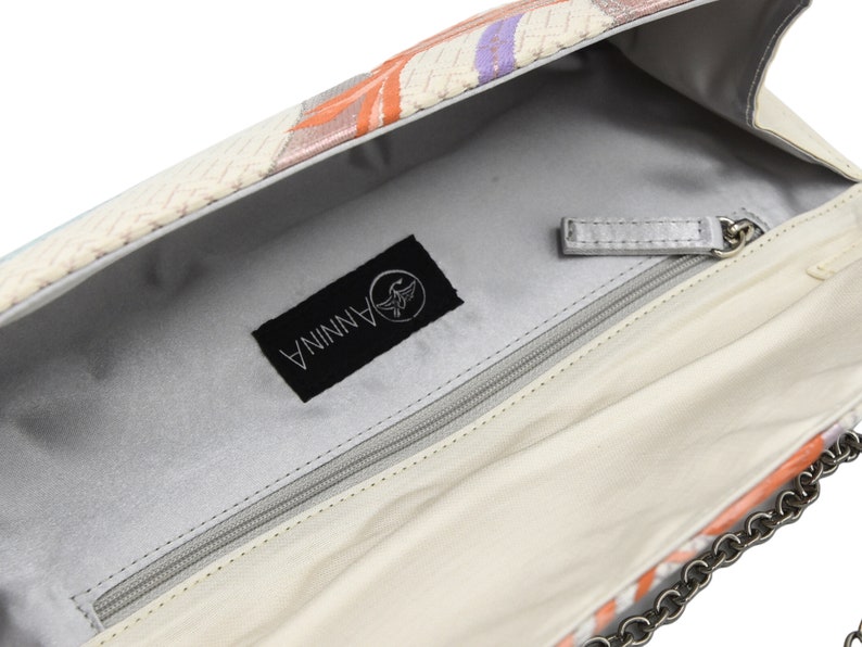 Puede incluir: El interior de un bolso de mano blanco con un bolsillo con cremallera y una correa de cadena plateada. El bolso tiene una etiqueta negra con la palabra "ANNINA" y un logotipo de p&aacute;jaro.