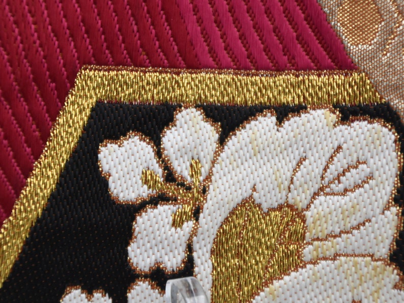 K&ouml;nnte beinhalten: Nahaufnahme eines schwarzen Stoffes mit goldenen und wei&szlig;en Stickereien. Die Stickerei zeigt ein florales Design mit wei&szlig;en Bl&uuml;tenbl&auml;ttern und goldenen Akzenten.