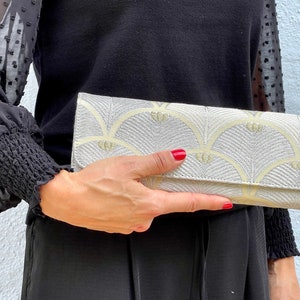 May include: A silver clutch bag with a yellow and white geometric pattern. The clutch has a flap closure and a silver zip.
