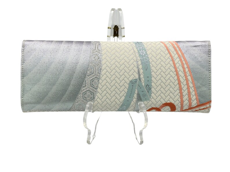 Puede incluir: Un bolso de mano plateado con un dise&ntilde;o estampado en blanco y azul. El patr&oacute;n presenta un dise&ntilde;o geom&eacute;trico con una textura tejida y un motivo floral azul y naranja.