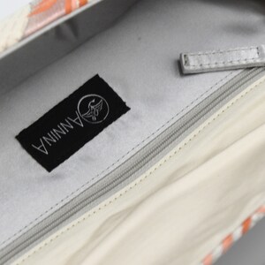 Puede incluir: El interior de un bolso blanco con una etiqueta negra que dice "ANNINA" con un logotipo de p&aacute;jaro. El bolso tiene una cremallera y un tirador de cremallera plateado.