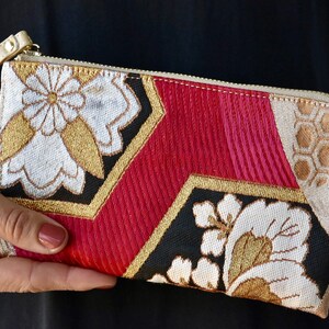 K&ouml;nnte beinhalten: Eine kleine, goldene, rote, schwarze und wei&szlig;e Stofftasche mit Rei&szlig;verschluss und einem goldenen Handgelenkband. Der Stoff hat ein Blumenmuster und die Tasche tr&auml;gt den Schriftzug "ANNINA" auf der Vorderseite.