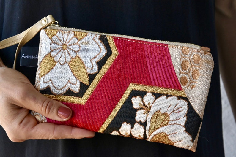 K&ouml;nnte beinhalten: Eine kleine, goldene, rote, schwarze und wei&szlig;e Stofftasche mit Rei&szlig;verschluss und einem goldenen Handgelenkband. Der Stoff hat ein Blumenmuster und die Tasche tr&auml;gt den Schriftzug "ANNINA" auf der Vorderseite.