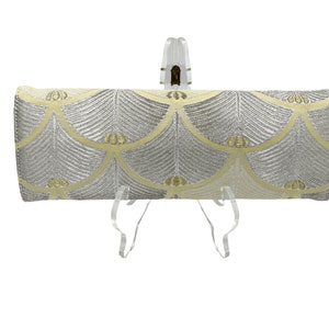 May include: A silver and yellow clutch bag with a geometric pattern. The bag is on a clear stand.