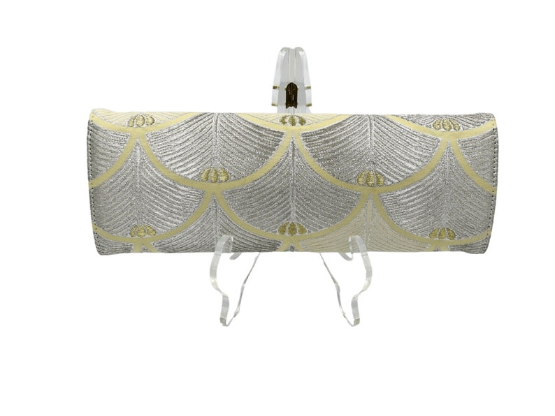 May include: A silver and yellow clutch bag with a geometric pattern. The bag is on a clear stand.