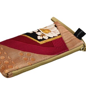 K&ouml;nnte beinhalten: Eine kleine, mit einem Rei&szlig;verschluss versehene Tasche mit einem beigefarbenen Stoff&auml;u&szlig;eren und einem bunten Patchwork-Design. Die Tasche hat ein rotes, goldenes und wei&szlig;es Blumenmuster auf schwarzem Hintergrund. Der Rei&szlig;verschluss ist golden und die Tasche hat einen schwarzen Lederriemen mit dem aufgedruckten Wort "ANNINA".