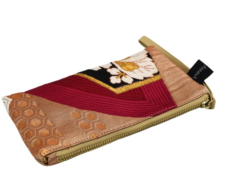 K&ouml;nnte beinhalten: Eine kleine, mit einem Rei&szlig;verschluss versehene Tasche mit einem beigefarbenen Stoff&auml;u&szlig;eren und einem bunten Patchwork-Design. Die Tasche hat ein rotes, goldenes und wei&szlig;es Blumenmuster auf schwarzem Hintergrund. Der Rei&szlig;verschluss ist golden und die Tasche hat einen schwarzen Lederriemen mit dem aufgedruckten Wort "ANNINA".