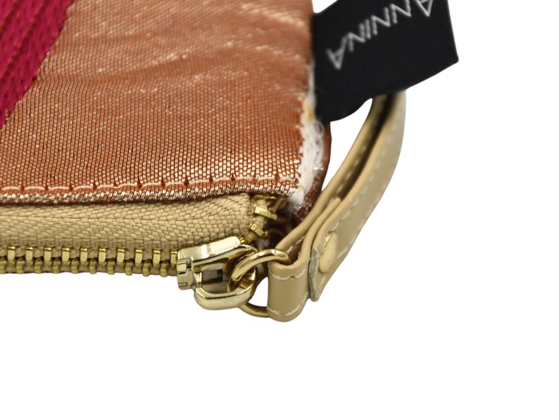 K&ouml;nnte beinhalten: Nahaufnahme eines goldenen Rei&szlig;verschlusses an einer beigefarbenen Stofftasche mit einem rosa-goldenen gestreiften Stofffutter. Die Tasche hat einen braunen Lederriemen mit einem Metallverschluss und ein schwarzes Etikett mit dem aufgedruckten Wort "ANNINA".
