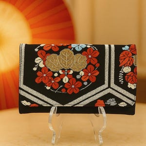 May include: Black clutch with floral embroidery in red, white, and blue. The design includes gold-colored leaves and silver accents. The clutch is displayed on a clear stand, with a red fan in the background.