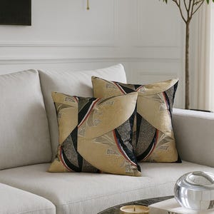Könnte beinhalten: Zwei dekorative quadratische Kissen mit einem geometrischen Muster in Gold, Schwarz, Rot und Weiß liegen auf einem hellen Sofa. Die Kissen haben eine strukturierte Oberfläche. Die Szene spielt in einem gut beleuchteten Wohnzimmer.