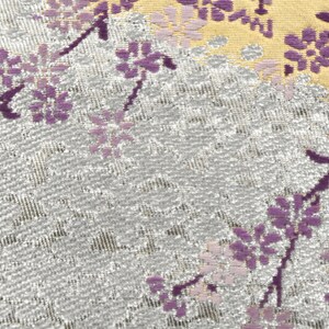 May include: A close-up of a silver fabric with a repeating pattern of purple flowers. The fabric has a subtle shimmer and a textured surface.