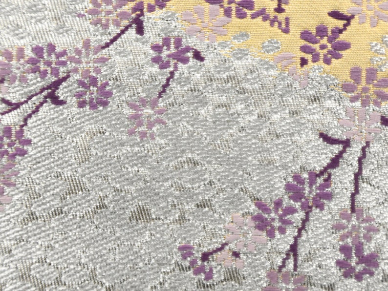 May include: A close-up of a silver fabric with a repeating pattern of purple flowers. The fabric has a subtle shimmer and a textured surface.