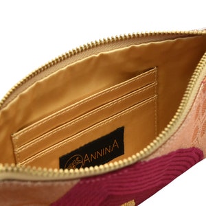 K&ouml;nnte beinhalten: Innenansicht einer Clutch aus rosa, rotem und beigefarbenem Stoff mit einem goldenen Rei&szlig;verschluss. Die Clutch hat drei innere Kartenf&auml;cher und ein schwarzes Leder-Etikett mit dem in Orange gedruckten Wort "ANNINA".