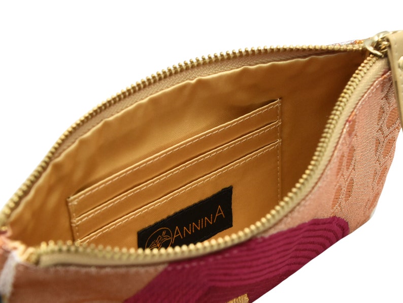 K&ouml;nnte beinhalten: Innenansicht einer Clutch aus rosa, rotem und beigefarbenem Stoff mit einem goldenen Rei&szlig;verschluss. Die Clutch hat drei innere Kartenf&auml;cher und ein schwarzes Leder-Etikett mit dem in Orange gedruckten Wort "ANNINA".