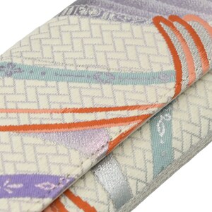 Puede incluir: Una cartera de tela blanca con un patr&oacute;n geom&eacute;trico en tonos naranja, morado y azul. La cartera tiene un brillo met&aacute;lico plateado y un borde doblado.