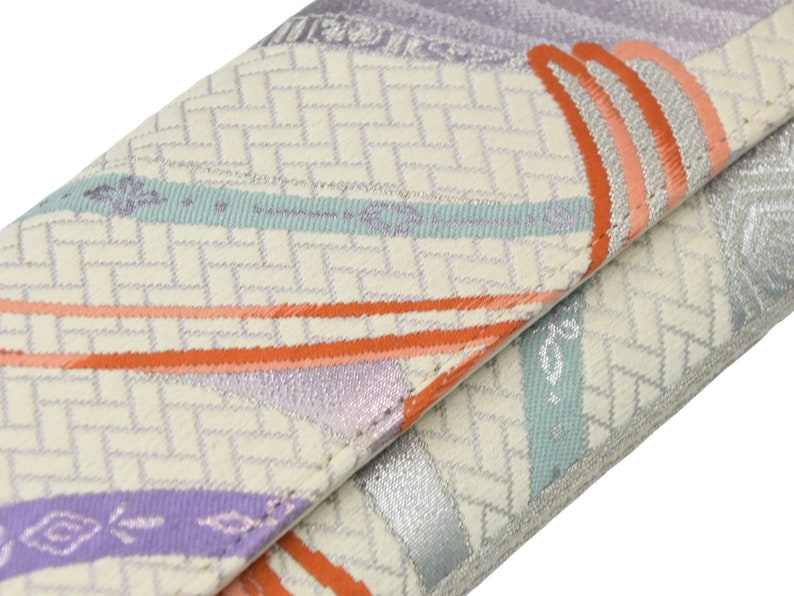 Puede incluir: Una cartera de tela blanca con un patr&oacute;n geom&eacute;trico en tonos naranja, morado y azul. La cartera tiene un brillo met&aacute;lico plateado y un borde doblado.