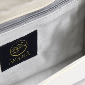 May include: The inside of a white handbag with a navy blue label that says "ANNINA" in gold lettering. The label has a gold image of a stylized bird.