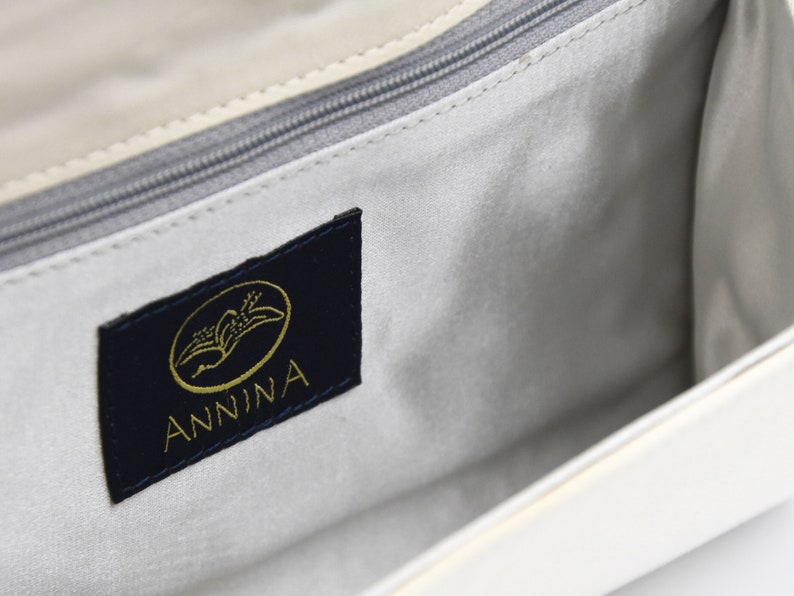 May include: The inside of a white handbag with a navy blue label that says "ANNINA" in gold lettering. The label has a gold image of a stylized bird.
