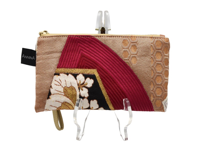 K&ouml;nnte beinhalten: Eine kleine, beige, rote und goldene Stofftasche mit Rei&szlig;verschluss und Blumenmuster. Die Tasche hat einen goldenen Rei&szlig;verschluss und ein kleines, schwarzes Etikett mit dem Wort "ANNINA" darauf.