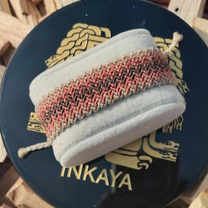 Könnte beinhalten: Ein gewebtes Armband mit einem braunen, beige und orangefarbenen Chevron-Muster. Das Armband wird auf einer schwarzen runden Box mit dem in goldenen Buchstaben geschriebenen Wort "INKAYA" präsentiert.