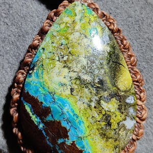 Peut inclure: Pendentif en forme de goutte d'eau avec une pierre vibrante, présentant des nuances de bleu, de vert, de jaune et de marron. La pierre est sertie dans un cordon marron tressé, créant un collier unique. Le pendentif est un bijou saisissant.