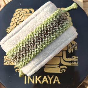 May include: A green and brown woven friendship bracelet with a chevron pattern. The bracelet is displayed on a small, beige, velvet box with the word "INKAYA" printed in gold on the front.