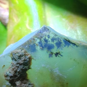 Op de afbeelding: Close-up van een doorschijnende, lichtgroene edelsteen met donkerblauwe en zwarte insluitsels. De steen heeft een ruw, getextureerd oppervlak met een lichtbruin en grijs gedeelte aan de zijkant. De achtergrond is een vage mix van groen en geel.