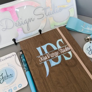 以下が含まれることがあります： 「Jolia Design Studio」という名前のパーソナライズされたビジネスブランディングアイテムのセットです。セットには、青と白のモノグラムが施された木製のジャーナル、会社のロゴが入った青いリボン、丸いロゴのキーホルダー、丸いロゴのマグネット式ブックマークが含まれています。