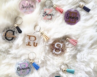 May include: Nine round clear glitter keyrings with different coloured glitter and tassels. Each keyring has a letter and a name on it. The names are Amelia, Zoe, Emma, Chloe, Raquel, Sophia, Brianna, and Juliana.