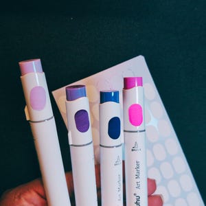 May include: Four brush tip markers with white barrels and coloured caps. The caps are lavender, purple, blue, and pink. The markers are labelled "Bruynzeel Art Marker".