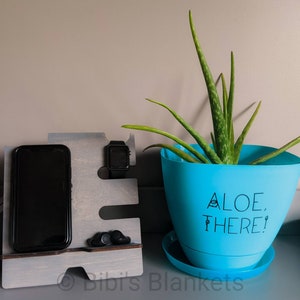 Könnte beinhalten: Ein grauer Holz-Telefonständer mit einem Telefon, einer Uhr und zwei Ohrstöpseln. Der Ständer steht auf einer grauen Oberfläche neben einem blauen Topf mit einer Aloe-Pflanze. Der Topf hat den Text "Aloe, There!" auf der Vorderseite.