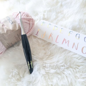 May include: A pink ball of yarn, a black crochet hook with a silver hook, and a white card with the letters of the alphabet in rainbow colors. The letters are B, C, D, E, F, G, G+, H, I, J, K, L, M, N, O, and P.