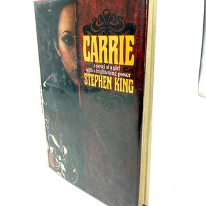 May include: A hardcover book titled Carrie by Stephen King, featuring a dark red and black cover with a portrait of a person on the left side. The text reads: Carrie, a novel of a girl with a frightening power, Stephen King, in yellow font.