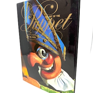 May include: A hardcover book titled "The Art of the Puppet" by Bil Baird. The cover features a colorful illustration of a puppet's face with a blue and gold hat, set against a black background. The book is displayed at an angle.