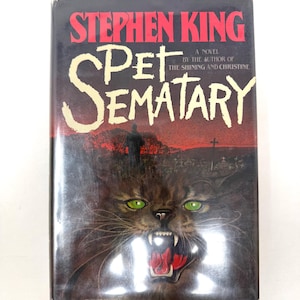 May include: A hardcover book titled Pet Sematary by Stephen King. The cover features a dark, red-toned graveyard scene with a silhouette of a person and a large, snarling brown cat with bright green eyes and sharp teeth in the foreground.