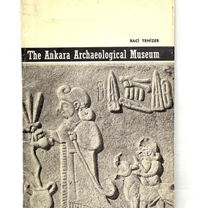 May include: A black and white photo of a book cover for "The Ankara Archaeological Museum" by Raci Temizer. The cover features a relief sculpture of two figures, one holding a staff and the other holding a small animal.