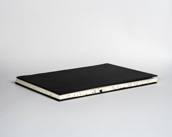 May include: A rectangular notebook with a solid black cover and cream-colored pages. The edges of the paper block feature a distinct black speckled pattern, creating a contrast against the dark cover. The notebook is positioned horizontally.